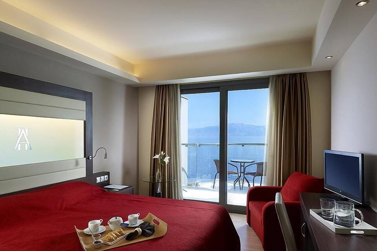 A hotel room with a large bed, a desk with a TV, a balcony with a table and chairs, and a view of the sea.