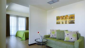 A hotel room with a green sofa, a small round table with a vase, a painting on the wall, and a bed with a green cover.