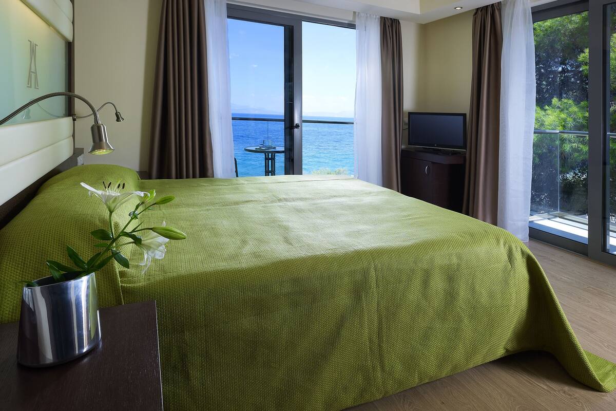 A hotel room with a bed, a desk, a TV, and a view of the sea.