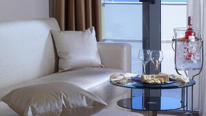 Habitación de hotel moderna con un sofá blanco, una mesa de vidrio con una botella y vasos, y vista al mar a través de una ventana.
