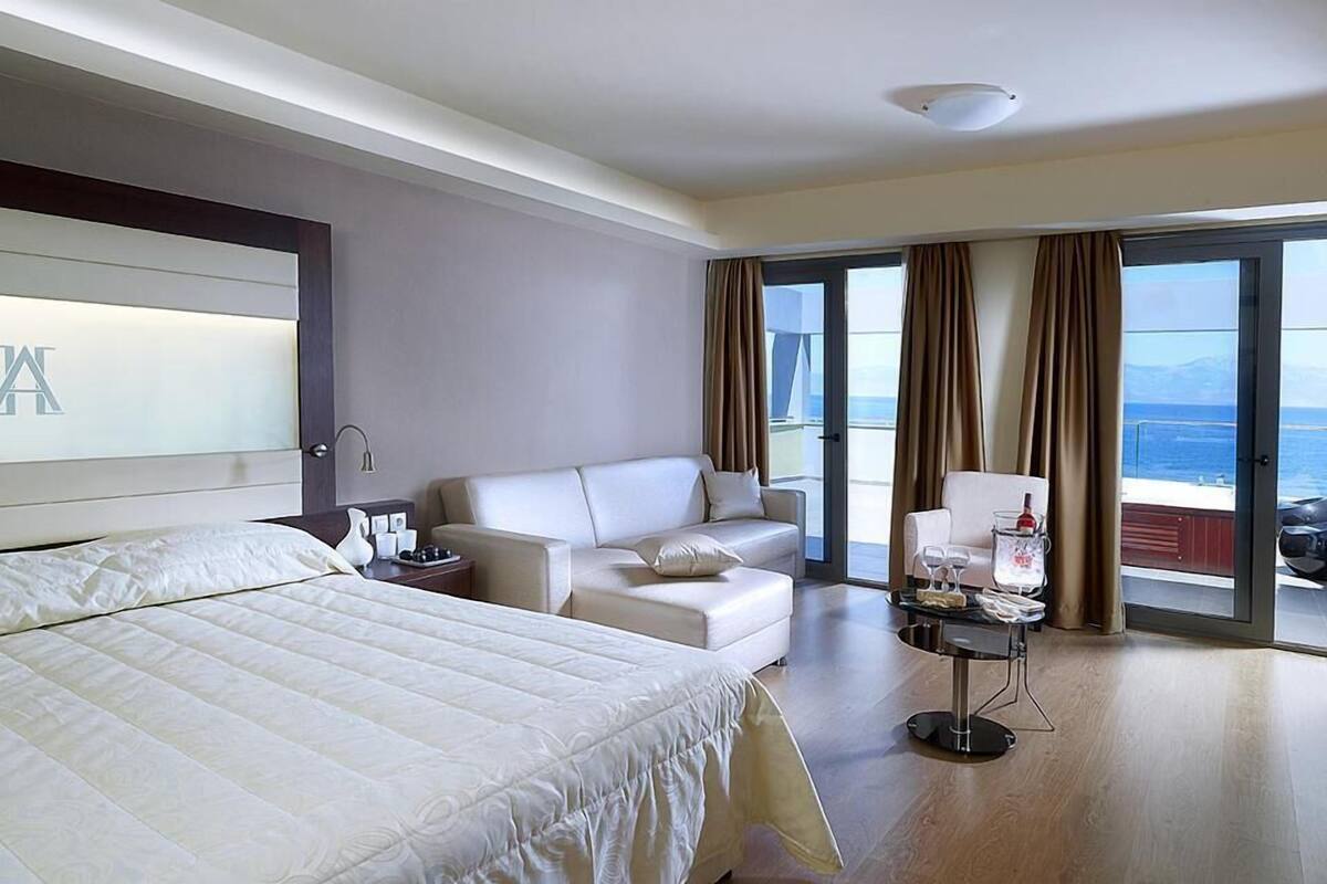 A hotel room with a bed, a sofa, a chair, a small table with a bottle, and a view of the sea.