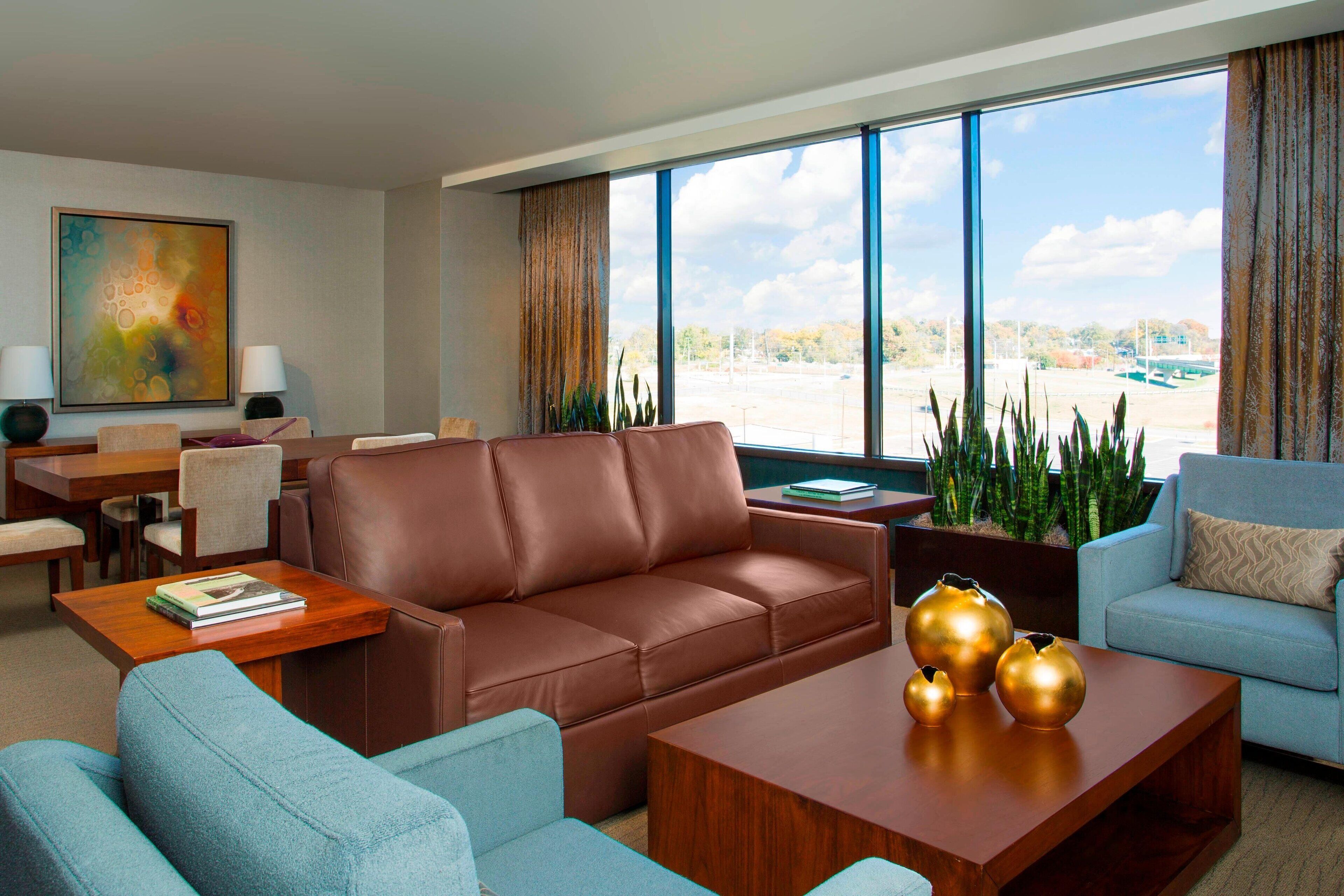 A modern living room with a brown leather sofa, a blue armchair, and a wooden coffee table with decorative vases. There is a dining area with a table and chairs, and a large window offering a view of the outside.