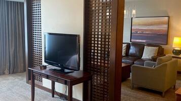 A hotel room with a TV on a wooden stand, a sofa, a chair, and a painting on the wall.
