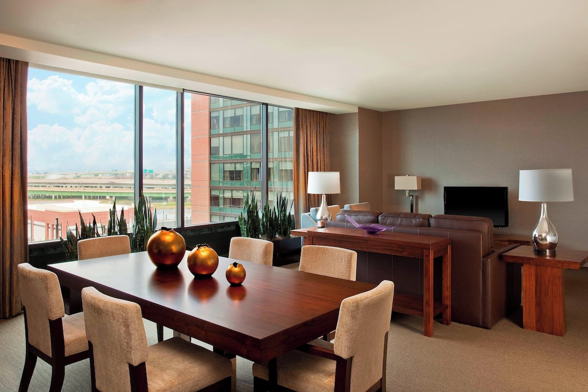 Habitación de hotel con comedor, sofá, televisor y vista al aeropuerto.