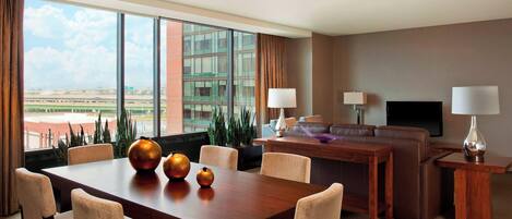 A hotel room with a dining area, a sofa, a TV, and a view of the airport.