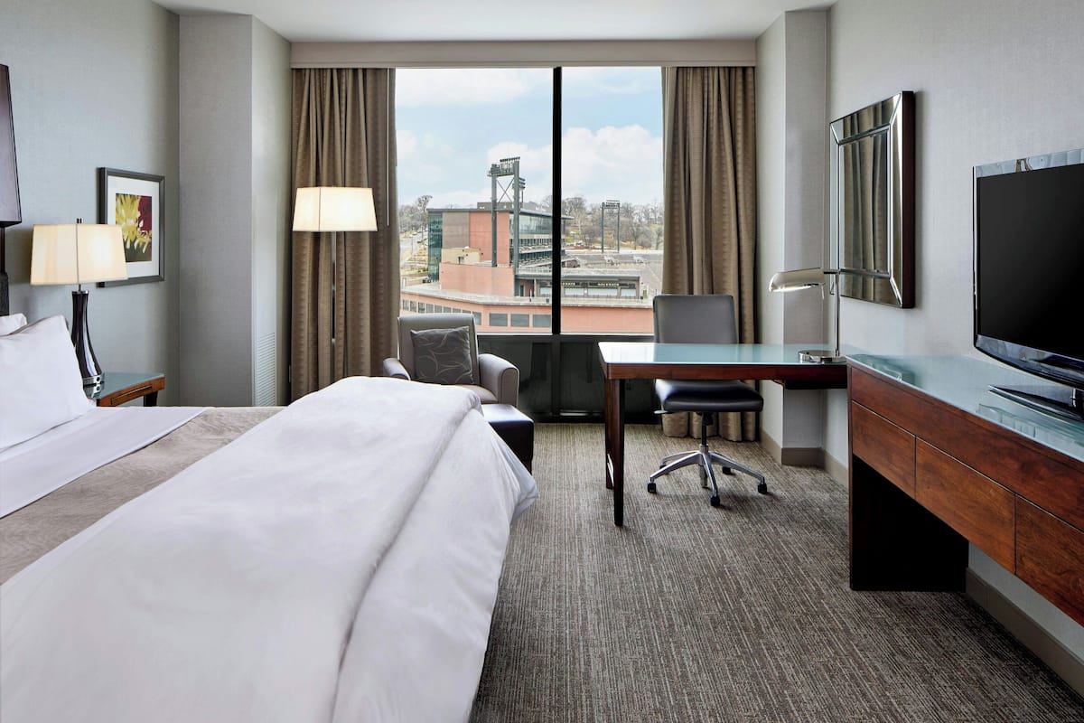 Habitación de hotel con una cama grande, un escritorio, una silla, un televisor y vista a un edificio a través de la ventana.