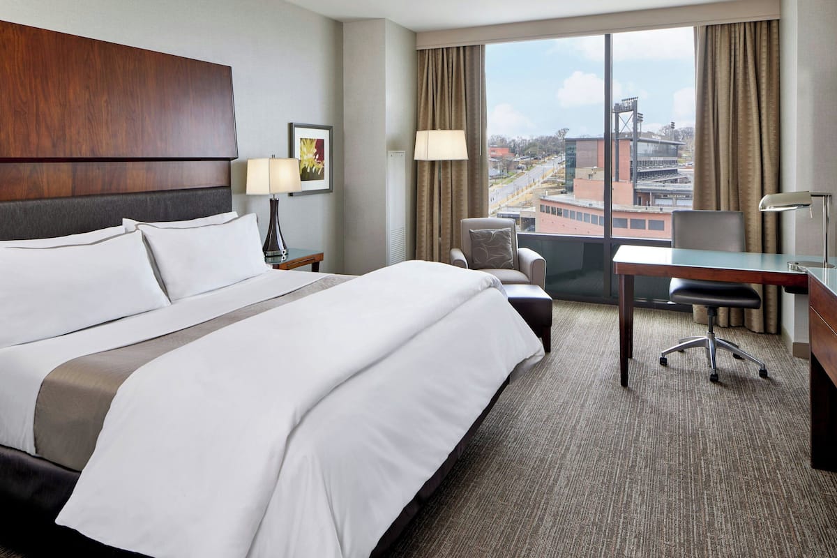 Habitación de hotel con una cama grande, un escritorio, una silla y vistas de la ciudad a través de grandes ventanales.