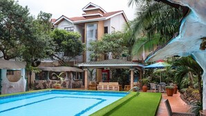 A swimming pool with a surrounding lawn, a covered seating area, and a building with a red-tiled roof.