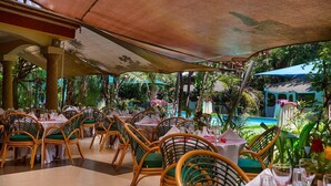 An outdoor restaurant with wicker chairs and tables, surrounded by lush greenery and a pool.