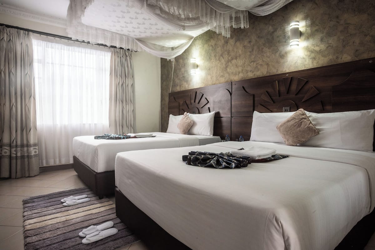 A hotel room with two beds, a large window with curtains, a headboard with a sunburst design, and a wall-mounted light fixture.