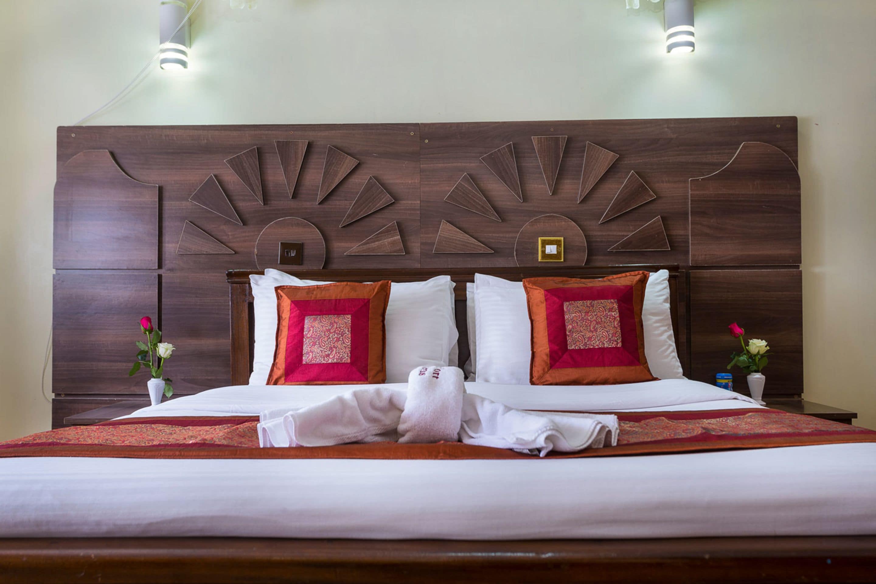 A neatly made bed with white linens, two red and one white pillow, and a decorative headboard.