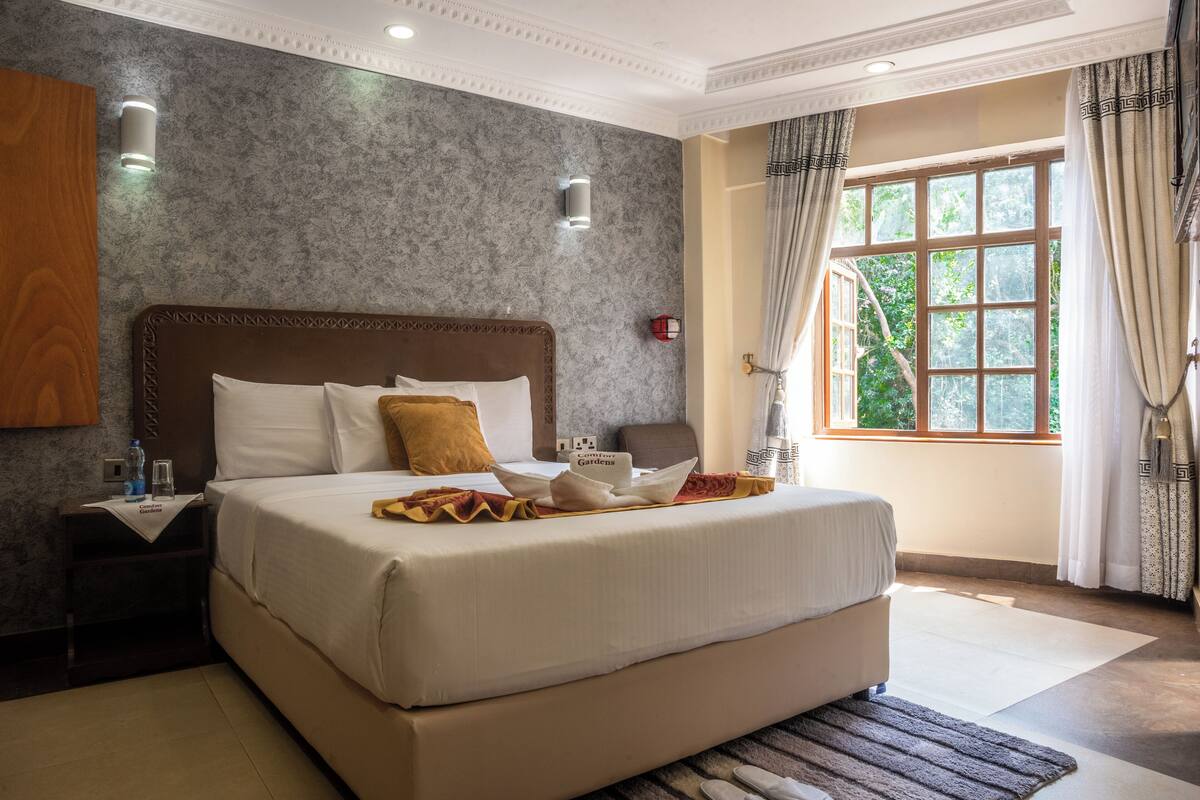 A large bed with a wooden headboard, white linens, and a decorative pillow. There is a nightstand with a glass of water and a bottle. A window with curtains and a view of greenery is visible.