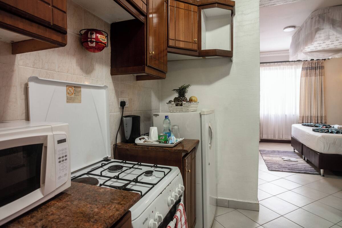A compact kitchen with a gas stove, microwave, and a small refrigerator. There is a basket of fruits on the counter and a bed visible in the adjacent room.