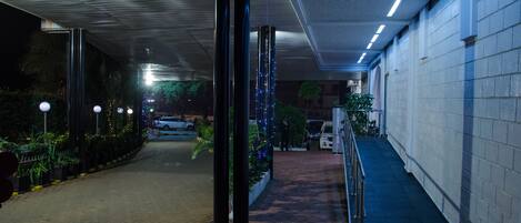 A covered walkway with a metal railing, potted plants, and a vehicle parked in the background.