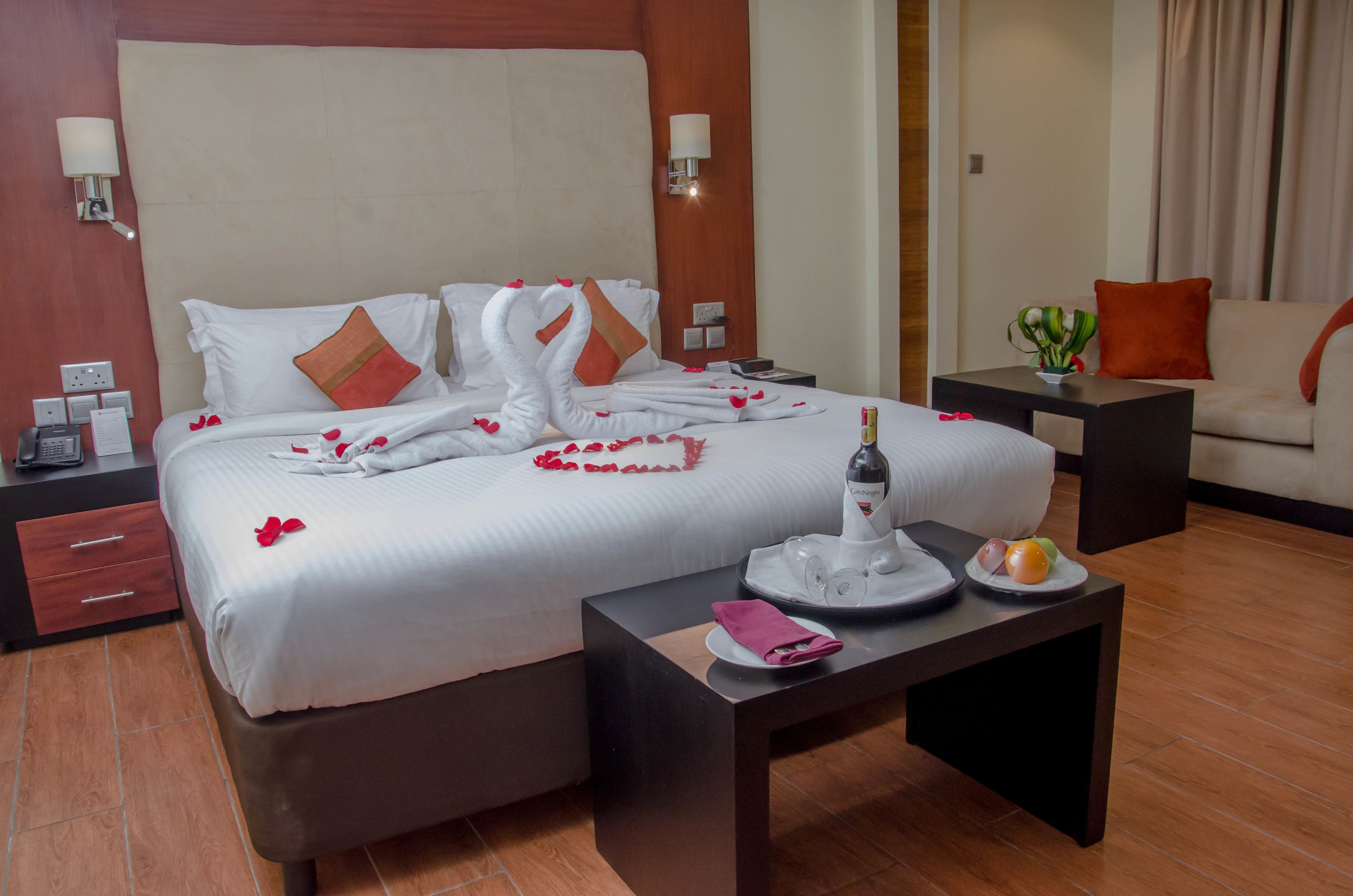 A hotel room with a large bed, a bedside table with a bottle and fruit, a small table with a plate and napkin, and a sofa with a cushion.