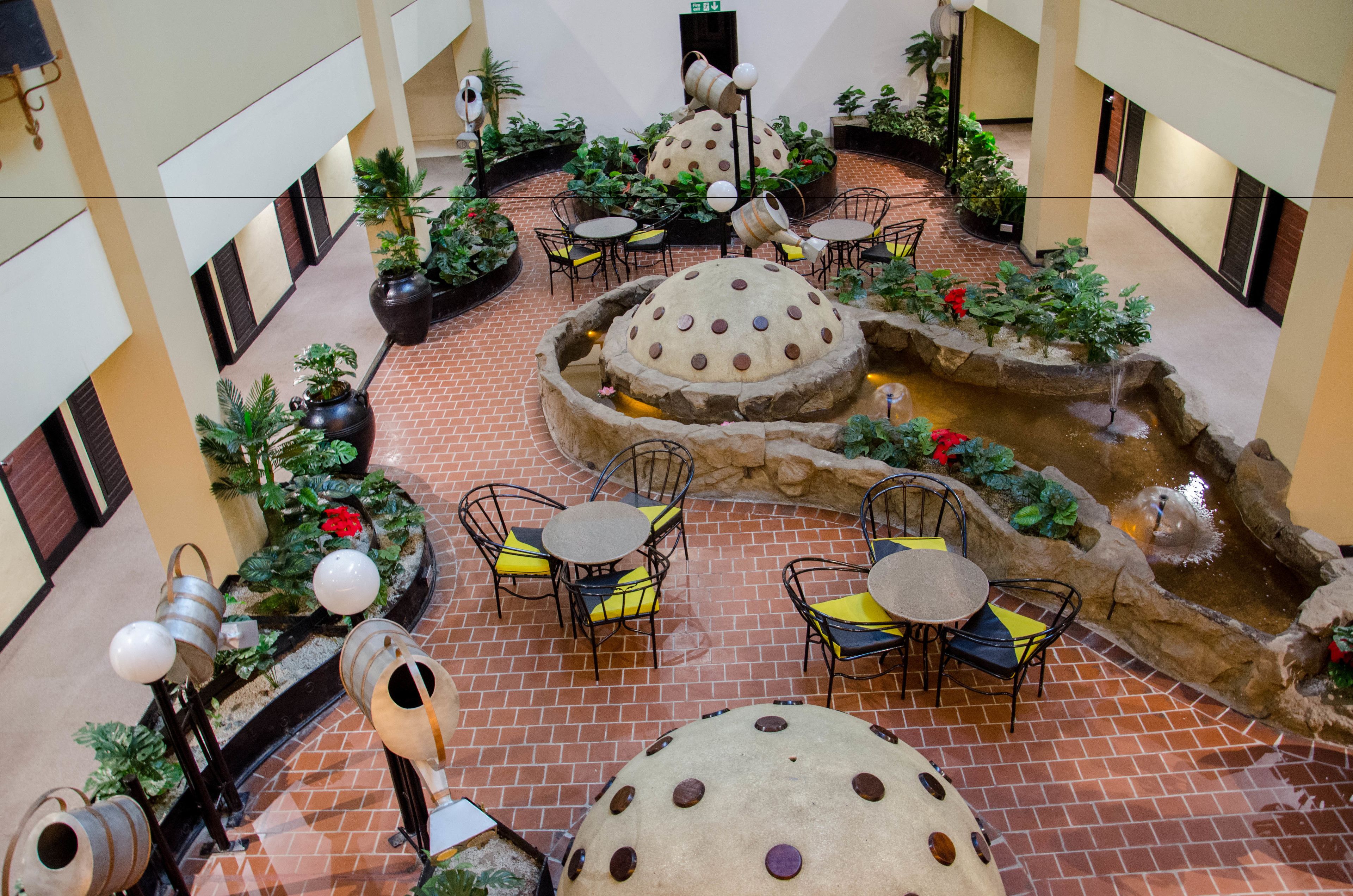 An indoor courtyard with brick flooring, multiple round tables and chairs, potted plants, and a central water feature.