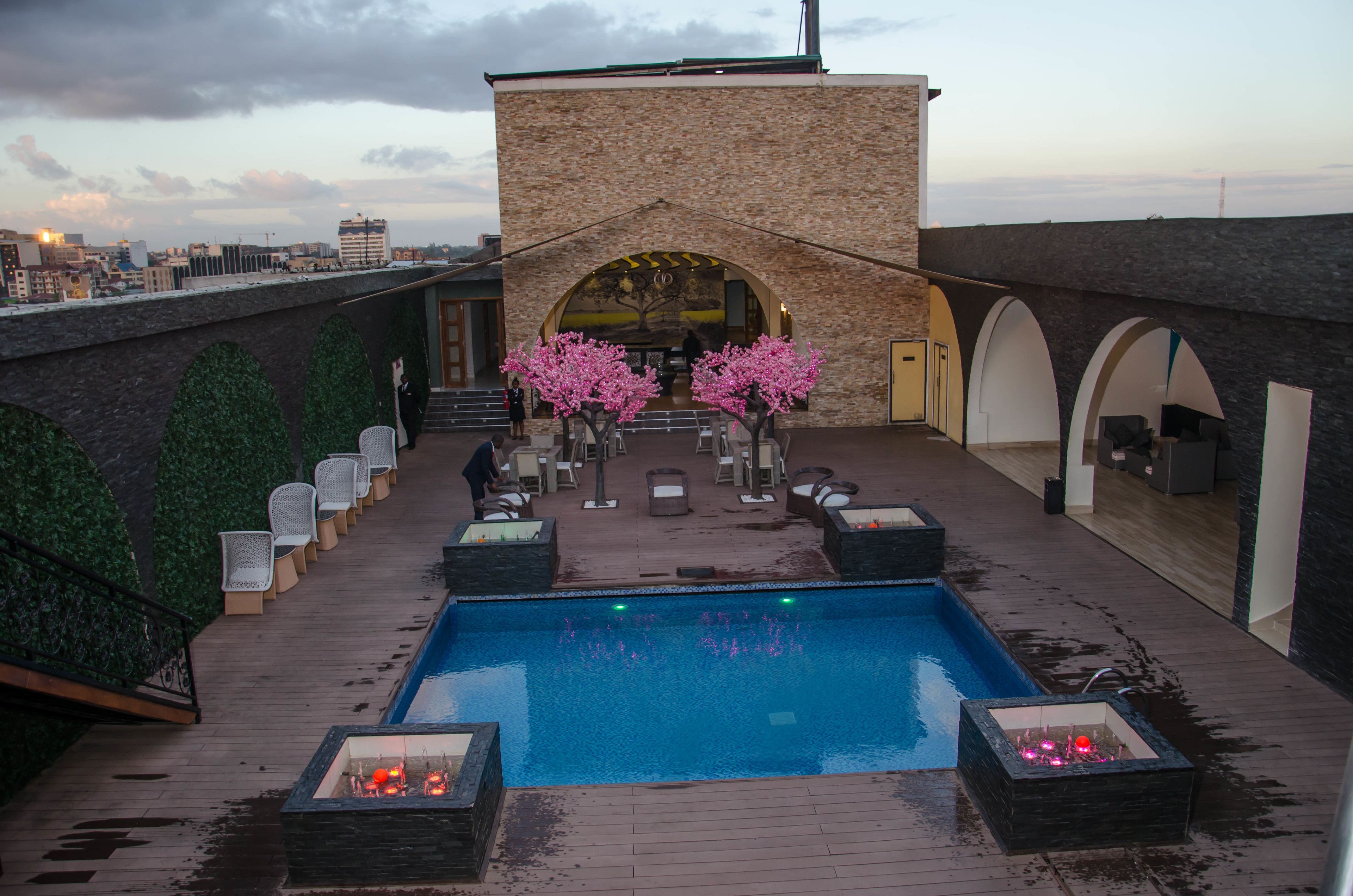 A rooftop pool area with a clear view of the city skyline, a stone building with an arched entrance, and a row of chairs along the pool's edge.