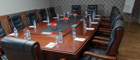 Un salón de conferencias con una larga mesa de madera, sillas de cuero negro, botellas de agua y jarrones con flores.