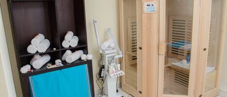 A room with a wooden cabinet filled with towels, a blue towel on the floor, a wooden sauna, and a massage machine.