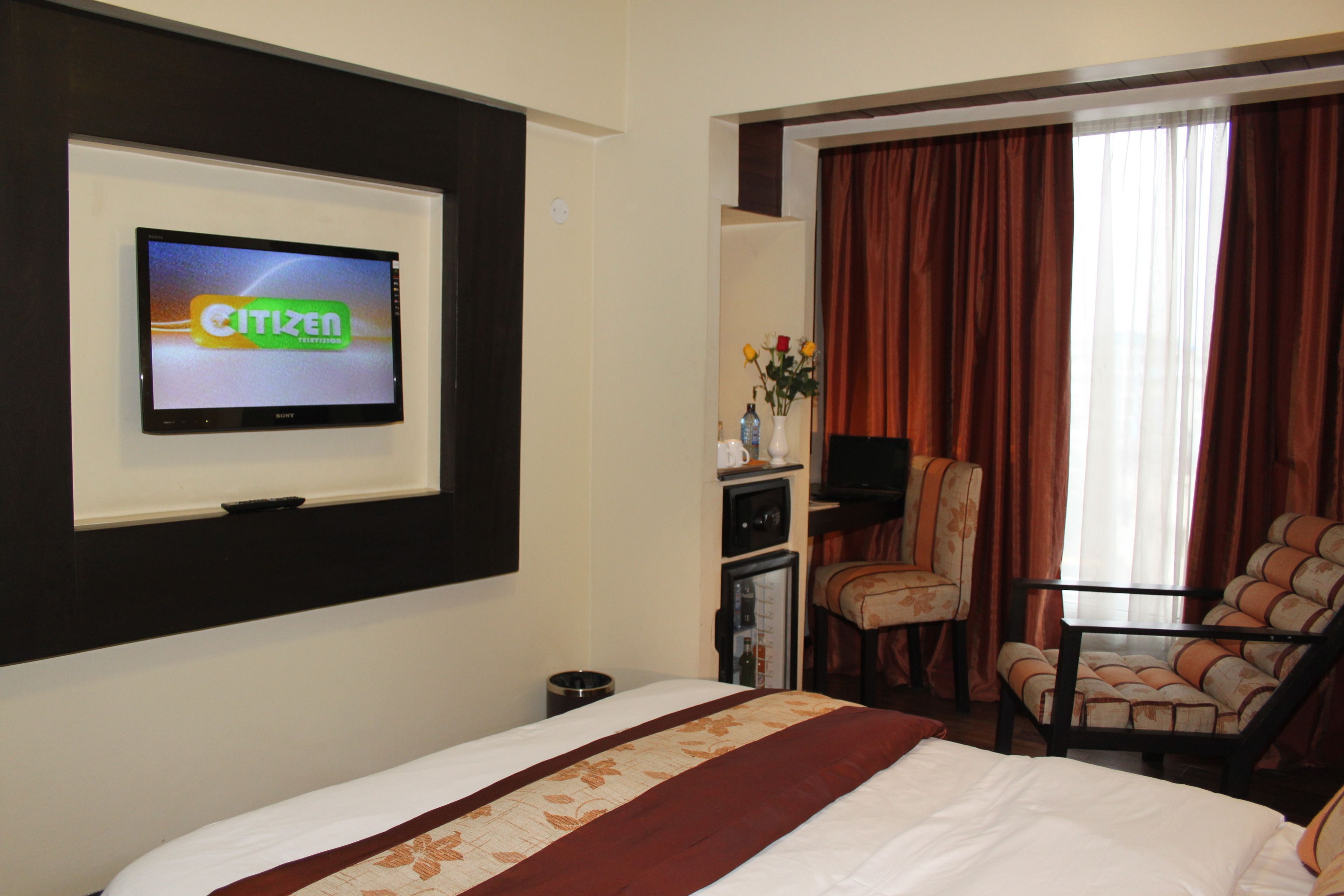 Habitación de hotel con cama, televisor montado en la pared, escritorio y silla, y ventana con cortinas.