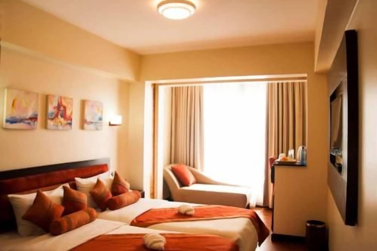 A hotel room with two beds, a sitting area, a large window with curtains, and artwork on the wall.