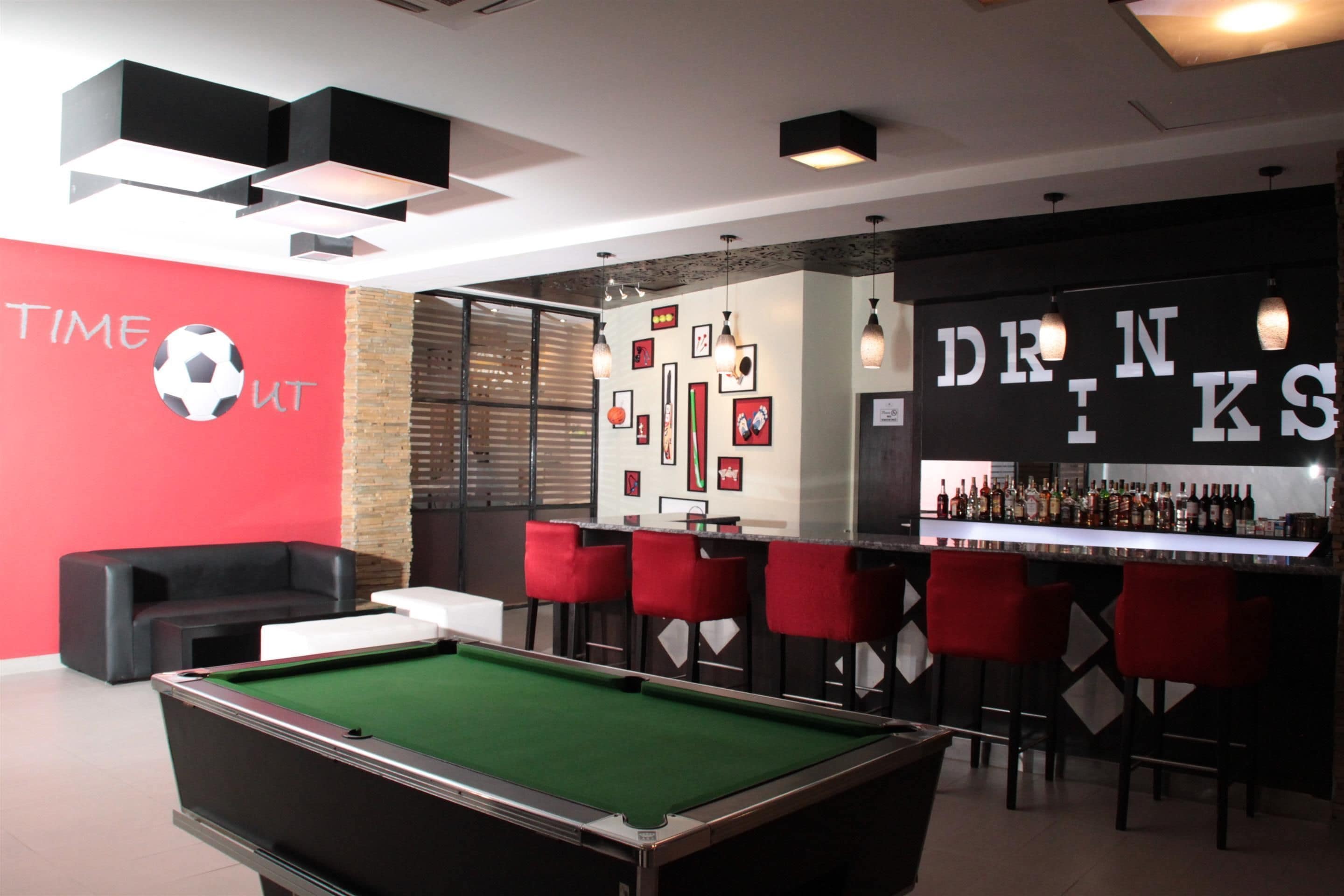 A pool table in a room with a red wall, a bar area with red chairs, and a wall with framed pictures and a soccer ball.
