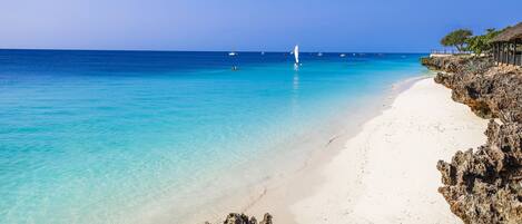 A beach with clear turquoise water, white sand, and rocky outcrops.