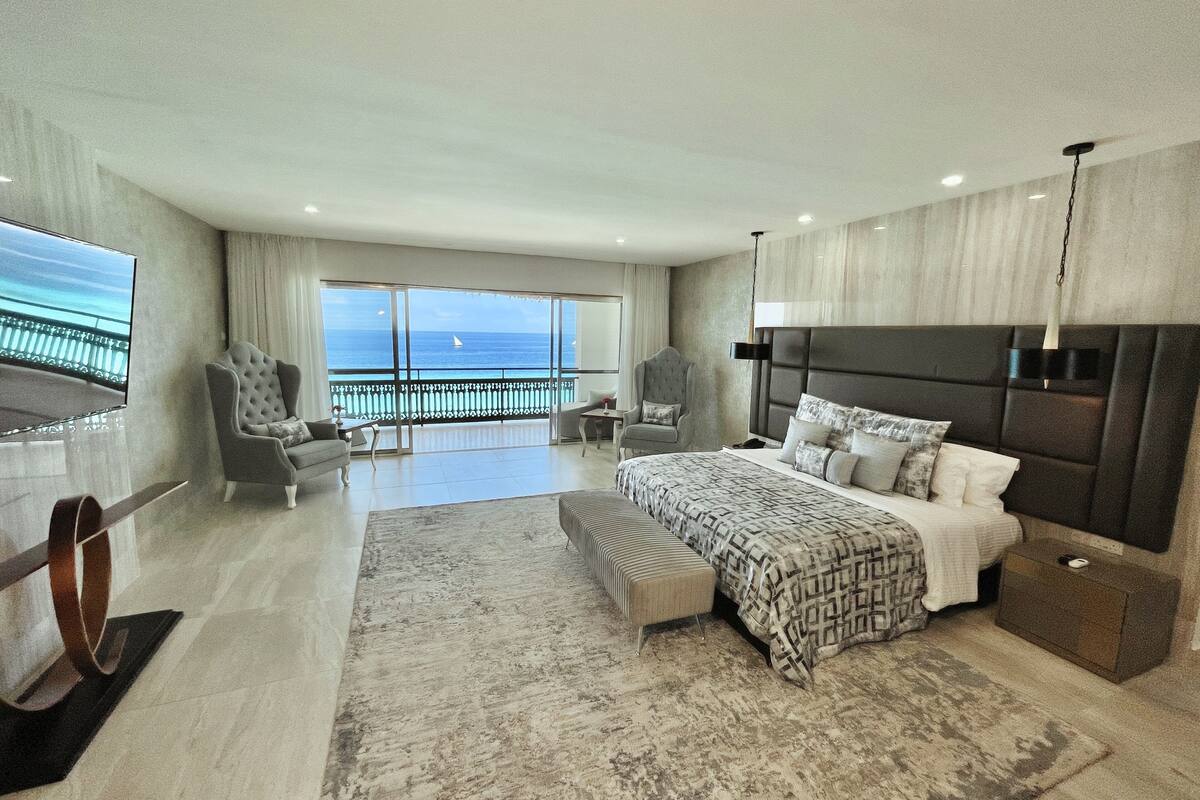 Un dormitorio amplio con una cama grande, un rincón de descanso con una silla y un sofá, televisión y un balcón con vistas al mar.