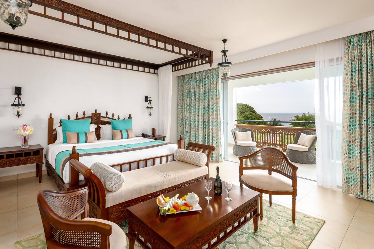 Habitación de hotel con cama, dos sillas, una mesa con fruta y copas de vino, y un balcón con vistas.