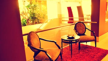 Una habitación con dos sillas, una mesita y un tapete rojo. Se aprecia una escalera al fondo y vistas al exterior con vegetación.
