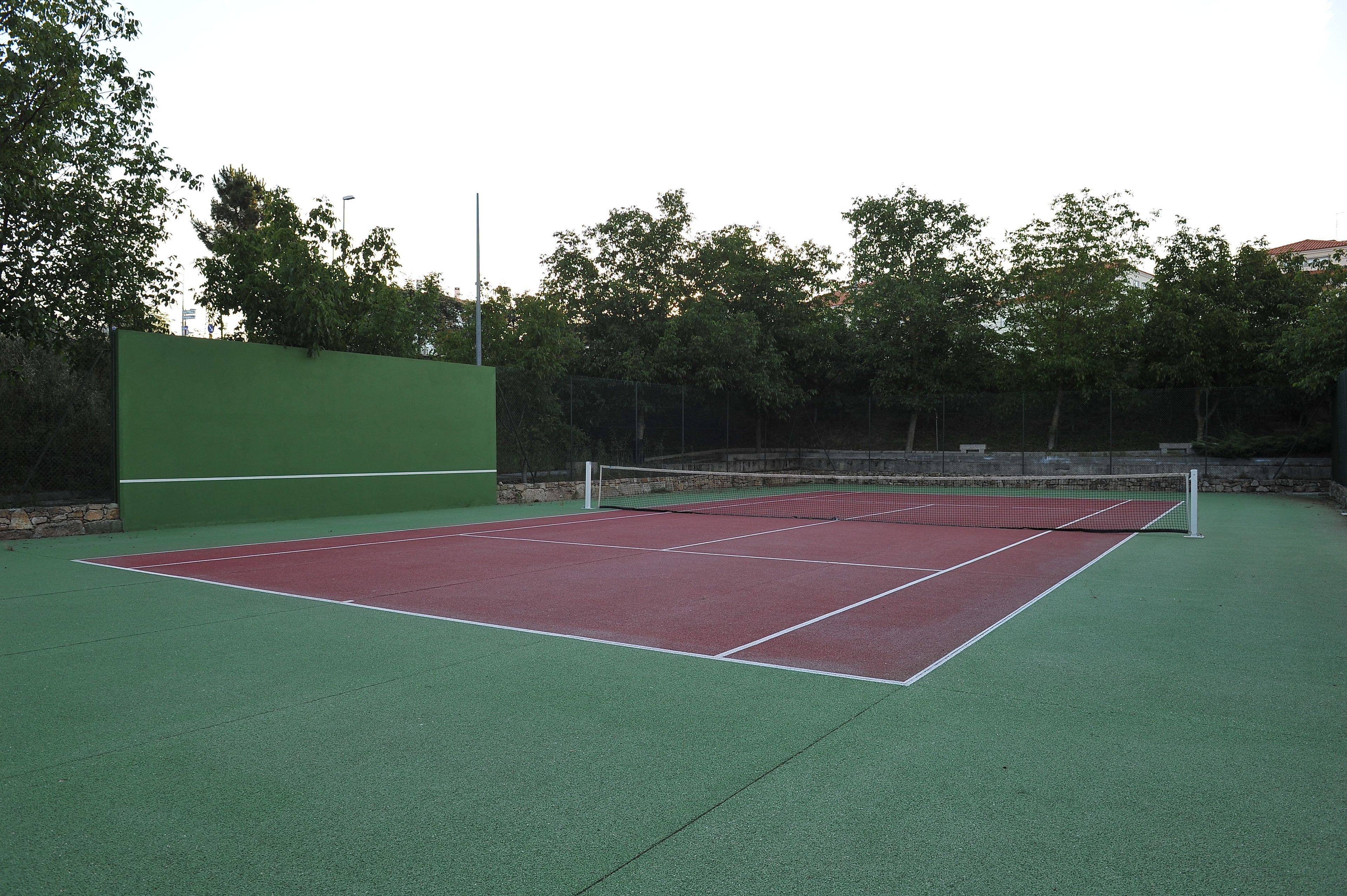 Una cancha de tenis con superficie verde y roja, rodeada de árboles y un muro de piedra.