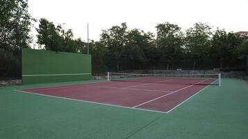 Una cancha de tenis con superficie verde y roja, rodeada de árboles y un muro de piedra.