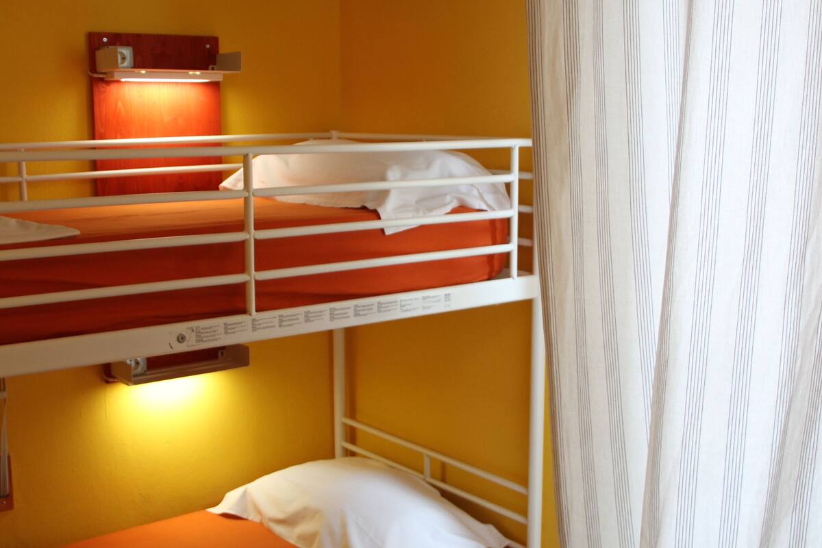 Ett rum med en våningssäng, orange sängkläder, en röd lampa och en randig gardin.