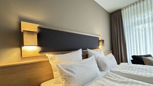 Een hotelkamer met een groot bed, een hoofdbord met ingebouwde verlichting en uitzicht op het omliggende landschap door dunne gordijnen.