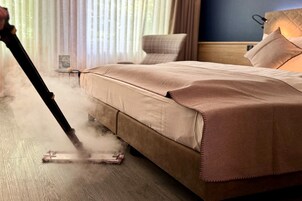 Una cama con cubierta color crema, un limpiador a vapor en el suelo y una ventana con cortinas.