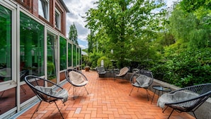 Een terras met bakstenen vloer, rieten stoelen en een gebouw met glazen wanden en een bakstenen gevel.