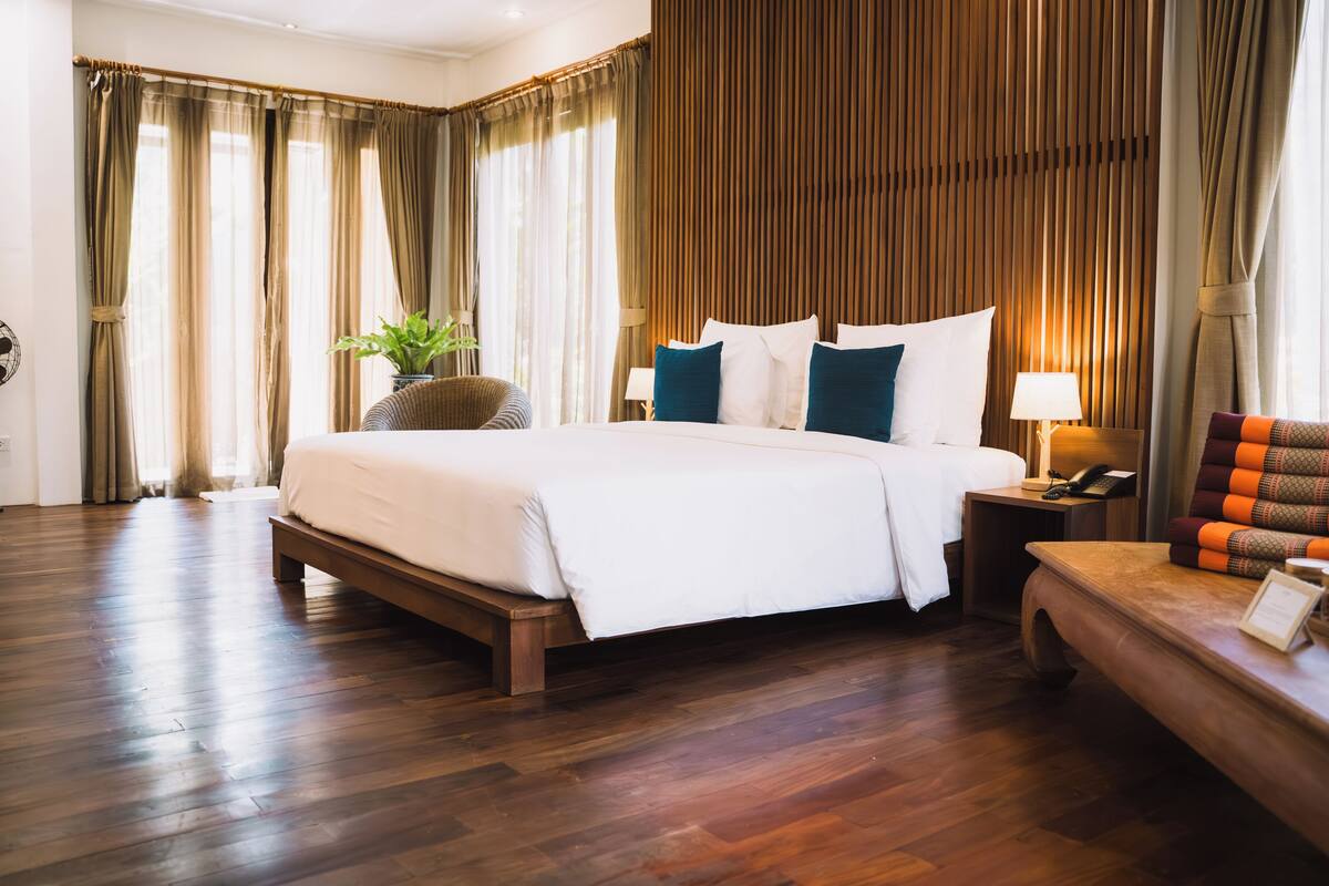 A hotel room with a large bed, wooden flooring, a bedside table with a lamp, and a view of the outdoors through curtains.