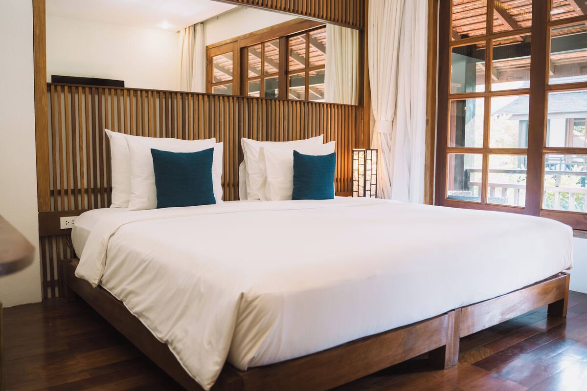 A large bed with white and teal bedding, wooden headboard, and a large window with a view of the outdoors.