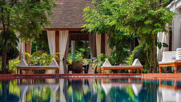 Uma área de piscina calma com reflexo, cercada por vegetação exuberante e um edifício tradicional com telhado em azulejos.