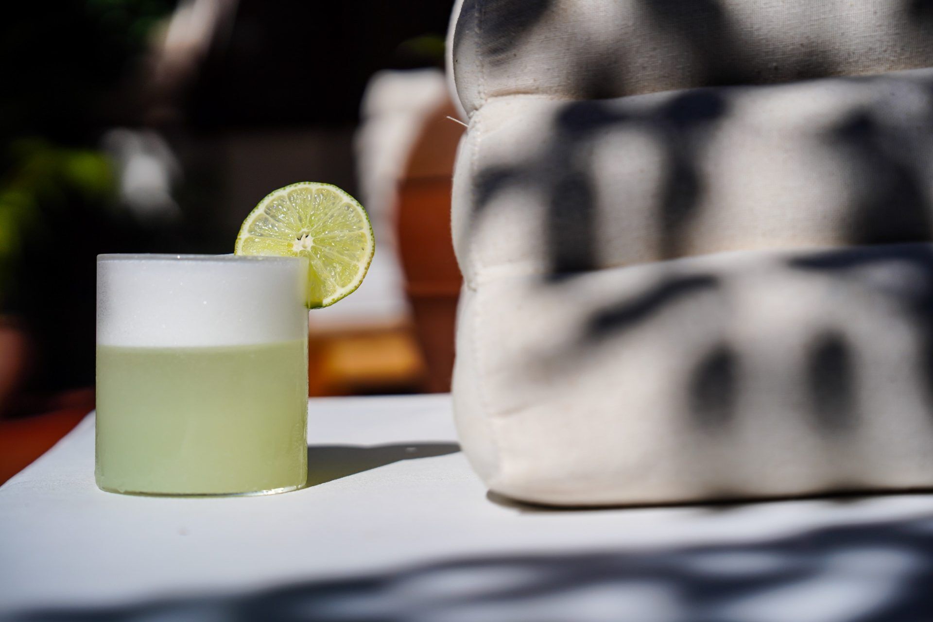 A glass of a light-colored beverage with a lime slice on a white surface, a patterned cushion, and a blurred background.