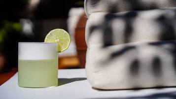 A glass of a light-colored beverage with a lime slice on a white surface, a patterned cushion, and a blurred background.