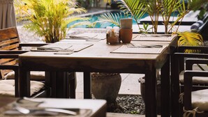 Uma área de refeições ao ar livre com mesa e cadeiras de madeira, cercada por vegetação exuberante e uma piscina.