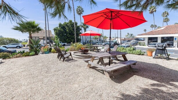 Een terras buiten met houten tafels en banken, rode parasols en een vuurkuil.