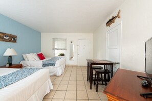 Habitación de hotel con dos camas, un escritorio y una silla. Se observa una ventana con persianas, una puerta y una pequeña mesa con lámpara.