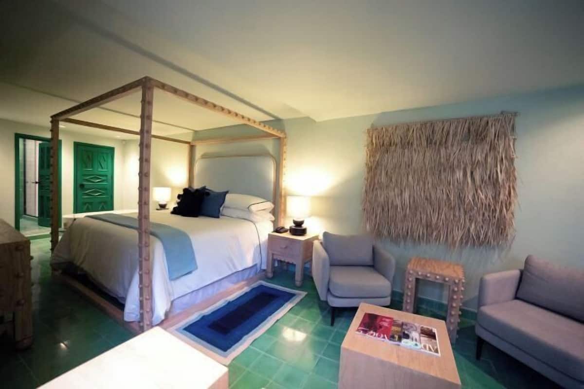 A four-poster bed, a small table, two chairs, a bedside table, and a woven wall hanging in a room with green flooring and a green door.