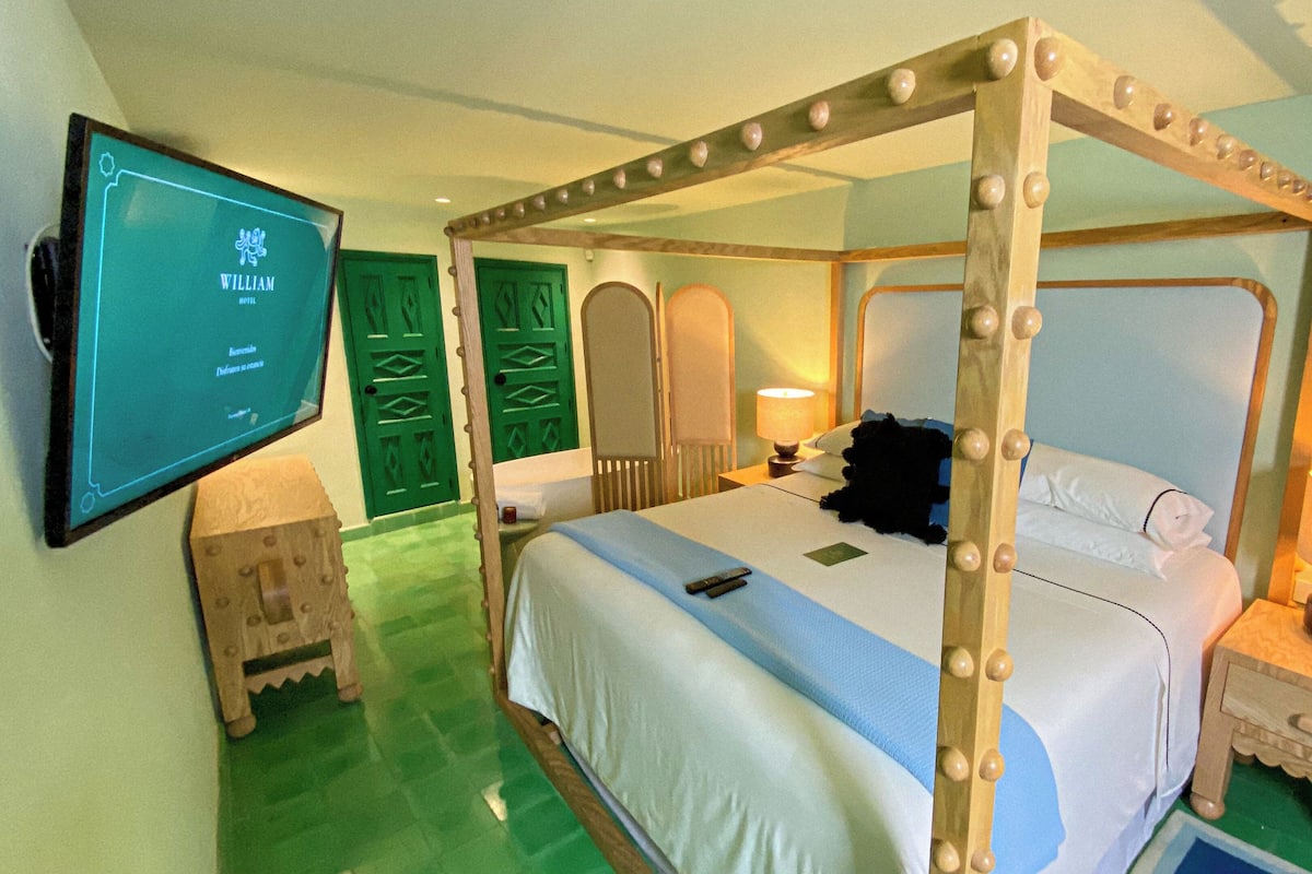 A four-poster bed with a television mounted on the wall, a bedside table with a lamp, and a small wooden cabinet.