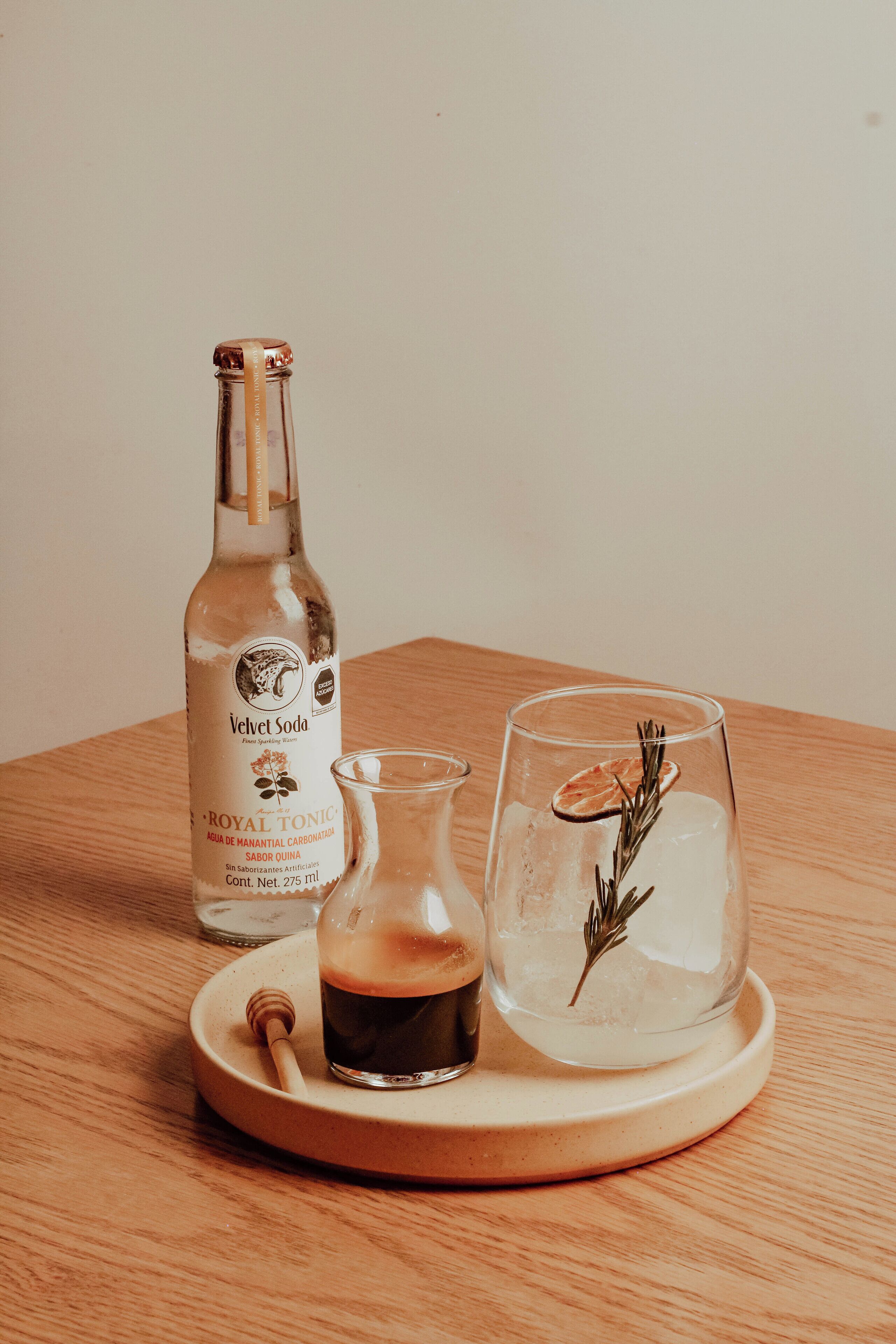 A bottle of Velvet Soda, a small glass pitcher, and a glass with a dried orange slice and rosemary on a wooden tray.
