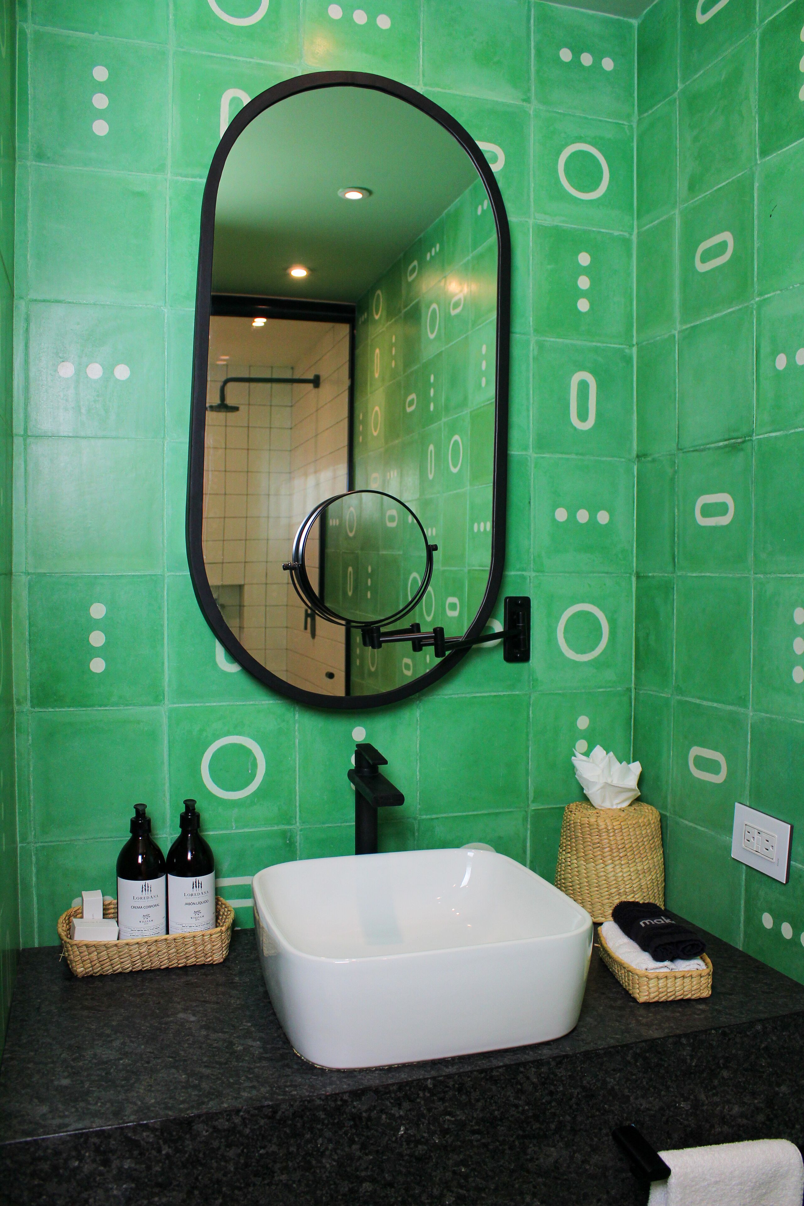 Une salle de bain avec des carreaux verts, un lavabo blanc, un miroir et des articles de toilette.