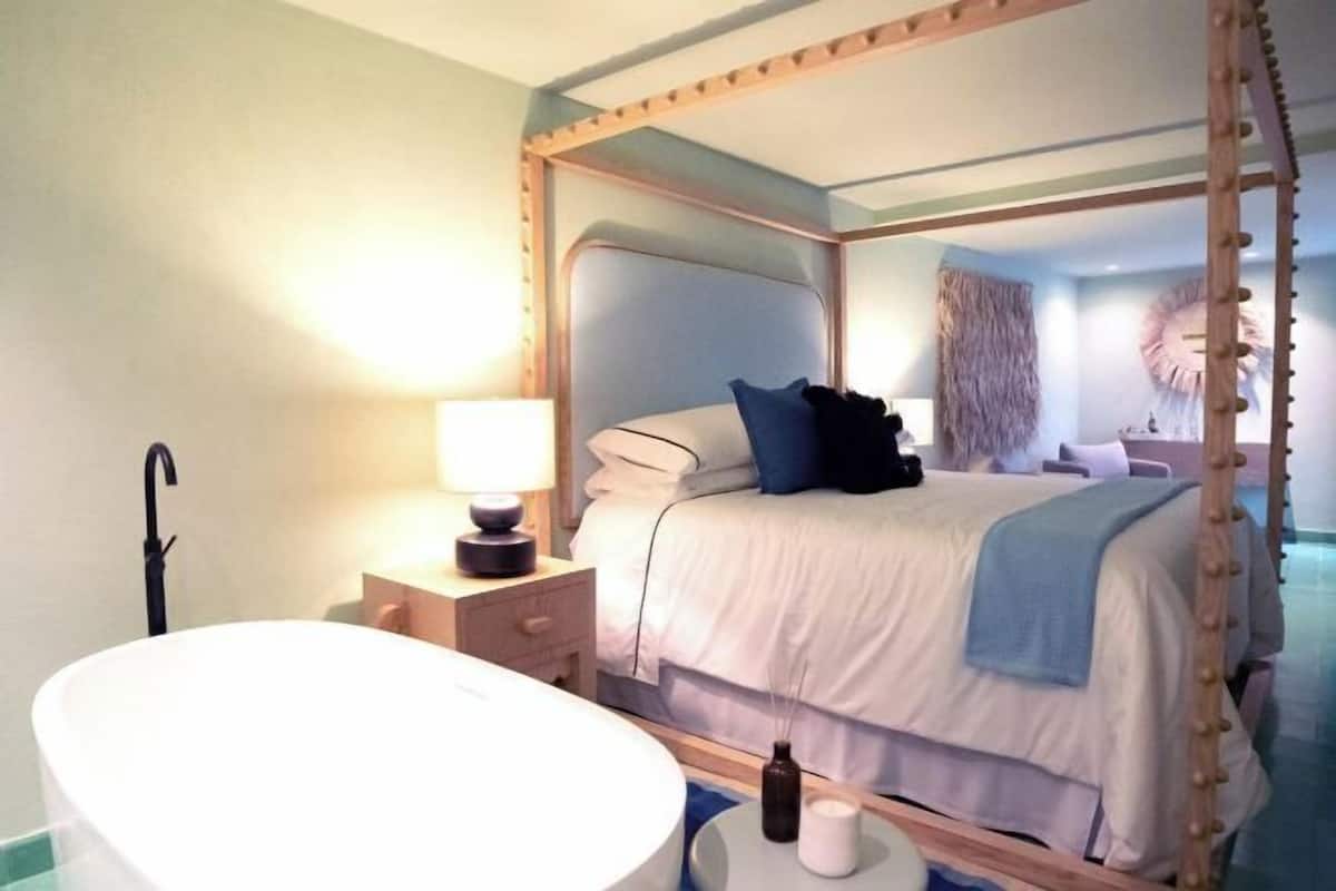 A four-poster bed with a white and blue color scheme, a round white bathtub, and a small bedside table with a lamp.