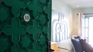 Una habitación de hotel con un diseño geométrico en la pared verde, una cama con ropa blanca, una mesita de noche y una lámpara.